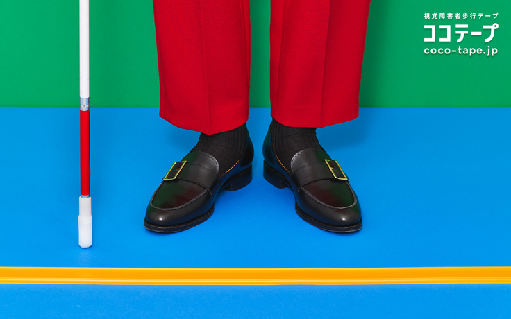 ココテープのビジュアル。背後の壁が緑、床は水色で黄色いココテープが横にまっすぐ引かれている。ココテープに両足を揃えて赤いズボン、黒いローファーを履き、白杖を持った人が立っている。