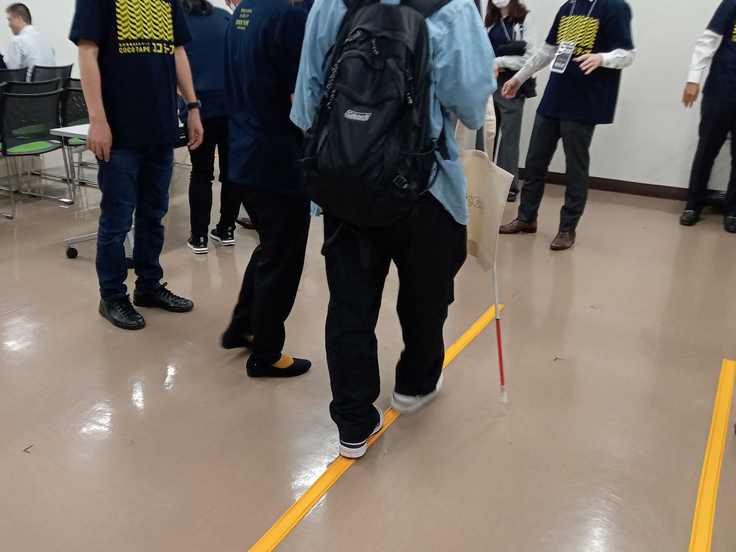 リュックを背負って白杖とトートバック持った人が床に設置されたココテープを歩行体験している写真。足でココテープを伝って歩いている。