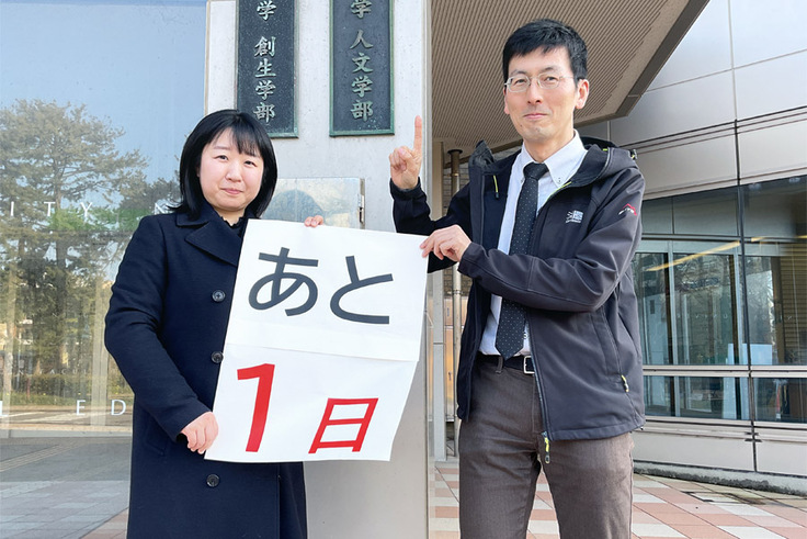 「人文学部」の看板の前で、「あと１日」と書かれた紙を持っているところの写真
