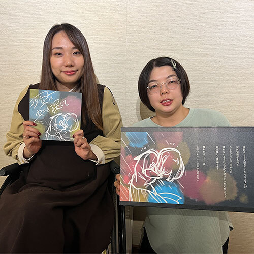 虹を探しての展示会で作成した絵本を持つ豆塚エリ（左）と、絵本の原画を持つこっちゃん（右）