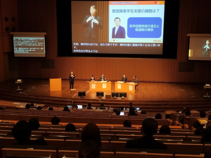 全体会の様子。ステージ中央に講師と司会が座っており、背後の大きなスクリーンにはスライド資料、手話通訳者（または手話話者の講師）、文字通訳の一部が合成されて映し出されている。ステージ下手には別途スクリーンがあり、文字通訳だけが映し出されている。その斜め前に手話通訳者。ステージ上手側にもスクリーンがあり、手話通訳者（または手話話者の講師）が映し出されている。