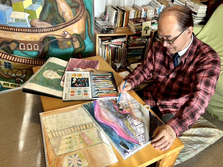 港座アートヴィレッジ代表・阿部剛がストーリーブックの原画を色鉛筆で書いている様子。机に絵や色鉛筆を広げて描いている様子を斜め上から撮影した画像。