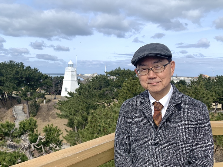 左奥に旧酒田灯台、右手前に港座アートヴィレッジ代表・阿部剛がたたずむ写真。