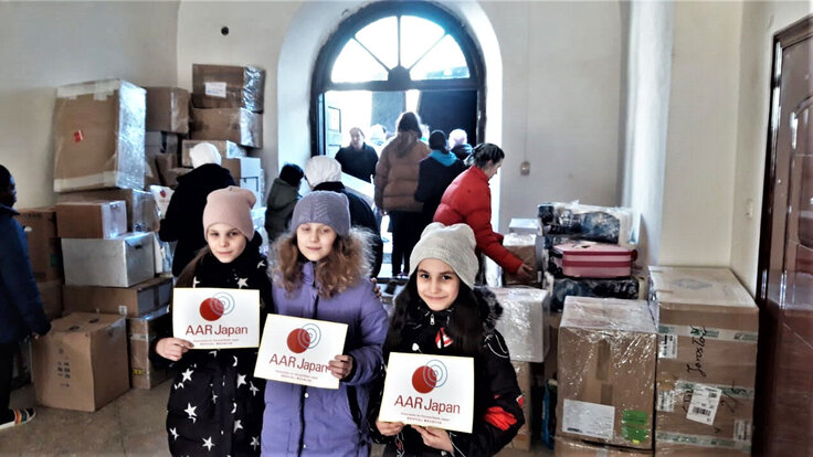 修道院に運び込まれた支援物資の前でAARのロゴを手にして笑顔の少女3人