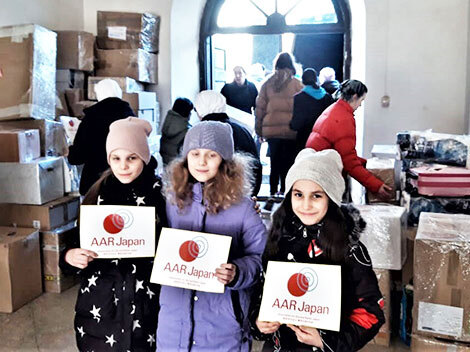 ウクライナ国内の避難民が身を寄せる修道院にAARから届いた荷物の前で笑顔の少女3名　手にはAARのステッカー